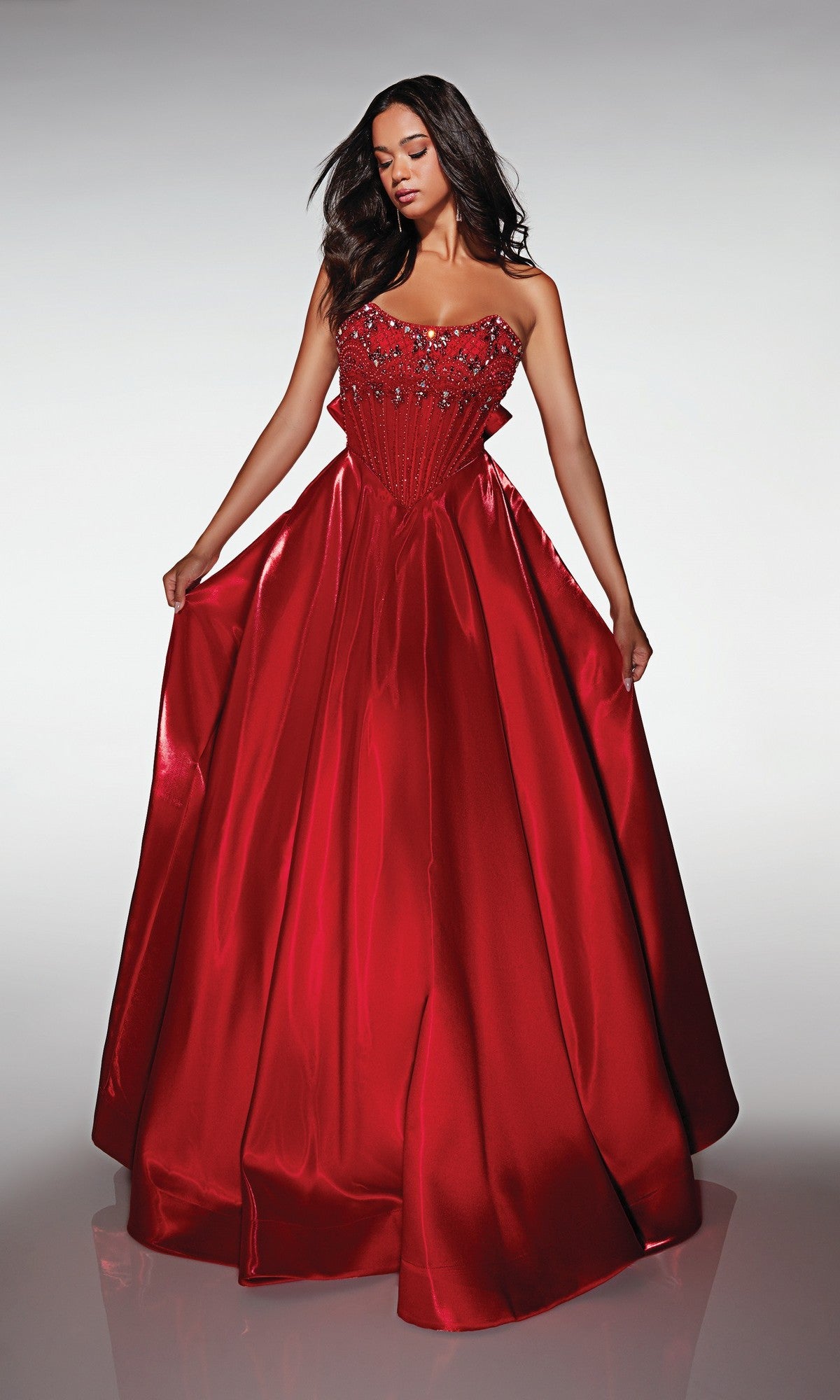 Wearing the Alyce 62134 long prom dress in shiny red, a woman poses against a plain background, holding the sides of her strapless satin ball gown with an embellished bodice and floor-length skirt.